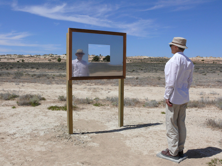 A man, who is standing side on to the camera, faces a rectangle mirror positioned on a frame. The mirror is set in a sandy, scrubby landscape and has a small rectangle cut out of it so you can see through to the view. You can see the man's reflection in the mirror.