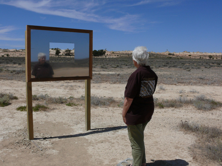 A woman, who is standing side on to the camera, faces a rectangle mirror positioned on a frame. The mirror is set in a sandy, scrubby landscape and has a small rectangle cut out of it so you can see through to the view. You can see the woman's reflection in the mirror.