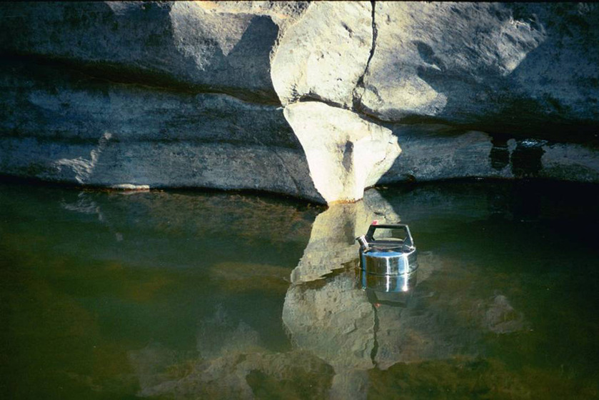 A photograph of a metal kettle floating in a river, large rocks standing along the river bank.