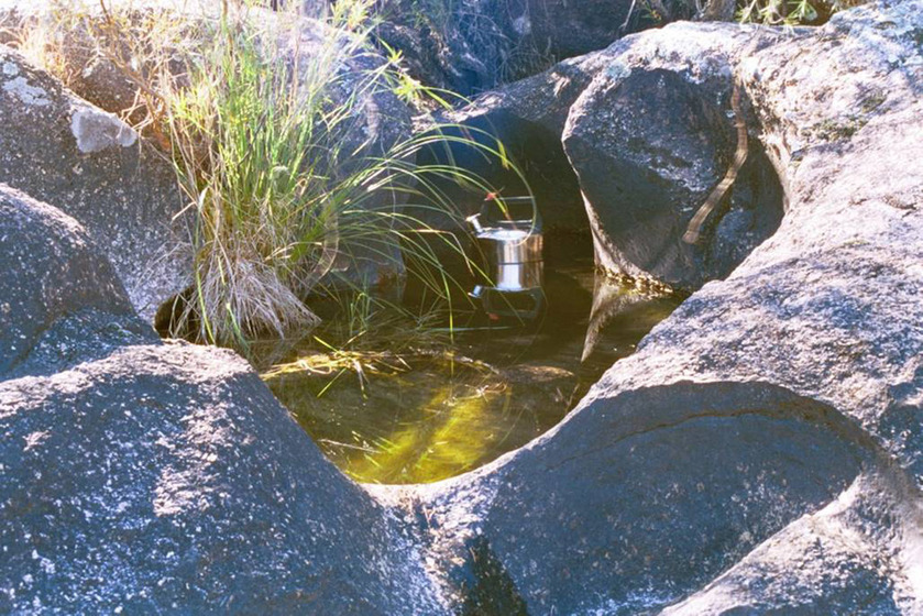 A photograph of a metal kettle floating in a river, large rocks positioned around the edge of the water.