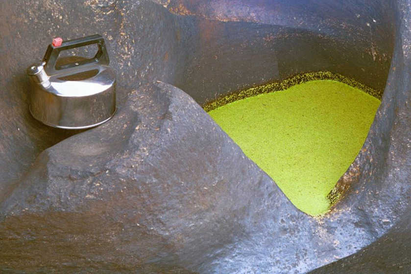 A photograph of a metal kettle positioned on large river rocks, a small puddle of water nearby a bright yellow colour.