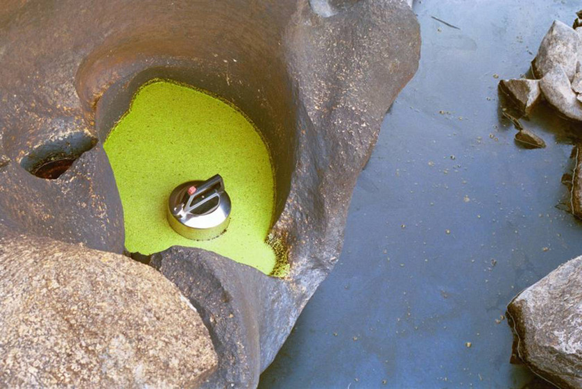 A photograph of a metal kettle positioned in a yellow puddle, large river rocks surrounding it and the river running just below.