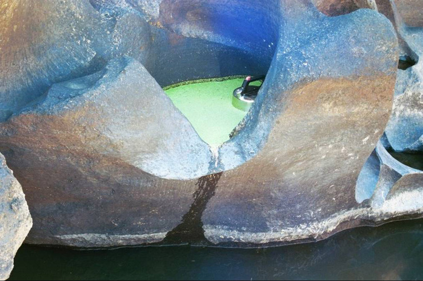 A photograph of a metal kettle positioned in a yellow puddle, large river rocks surrounding it and the river running just below.