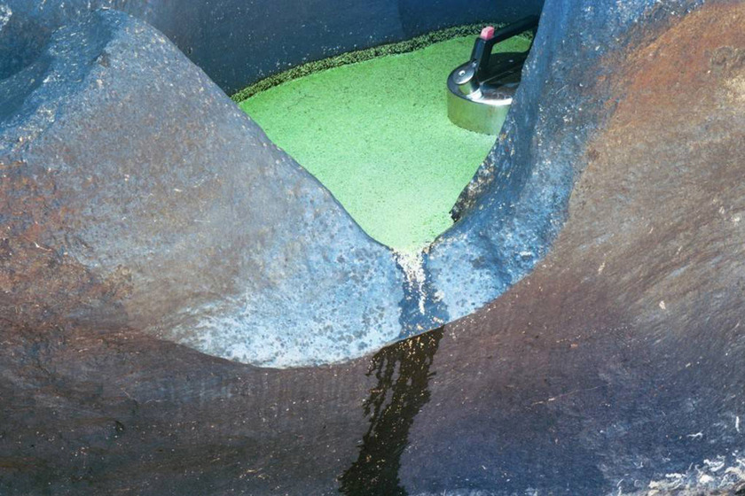 A photograph of a metal kettle positioned in a yellow puddle, large river rocks surrounding it and the slight trickle of water rishing over the edge.