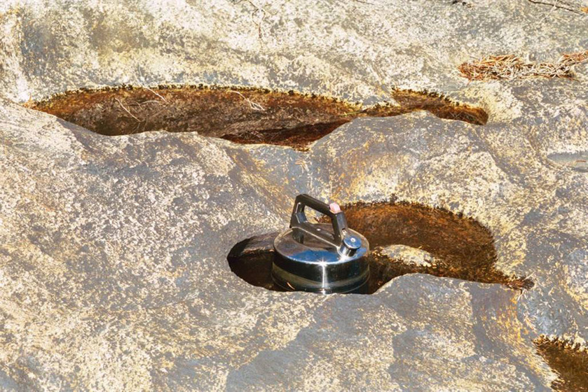 A photograph of a metal kettle positioned in a very small puddle of water, surrounded by river rocks and nearby shallow puddles.