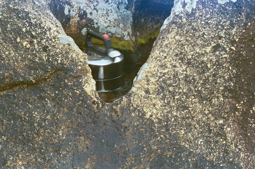 A photograph of a metal kettle positioned in a very small puddle of water, surrounded by river rocks and nearby shallow puddles.