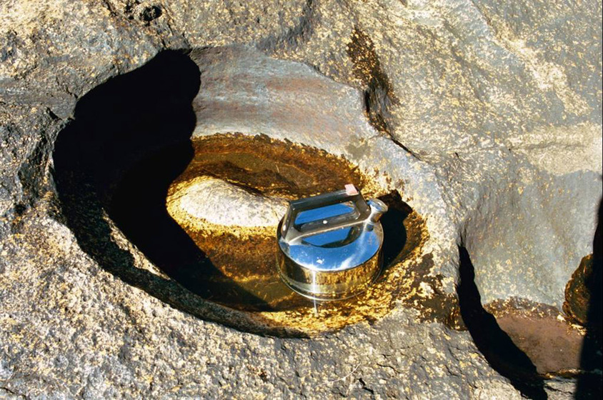 A photograph of a metal kettle positioned in a very small puddle of water, surrounded by river rocks and nearby shallow puddles.