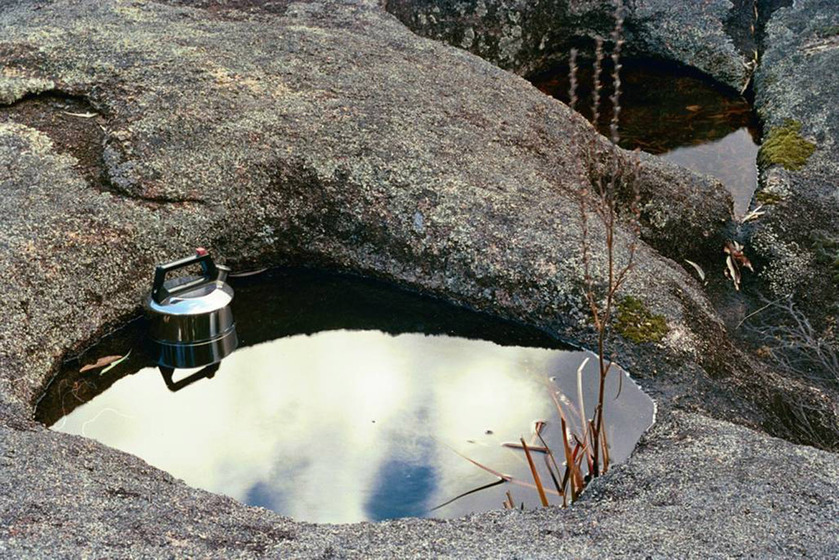 A photograph of a metal kettle positioned in a very small puddle of water, surrounded by river rocks and nearby shallow puddles.
