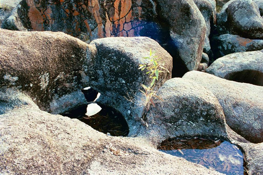 A photograph of a metal kettle positioned in a very small puddle of water, surrounded by river rocks and nearby shallow puddles.