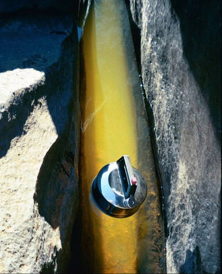 A photograph of a metal kettle floating in a river, large rocks positioned around the edge of the water.