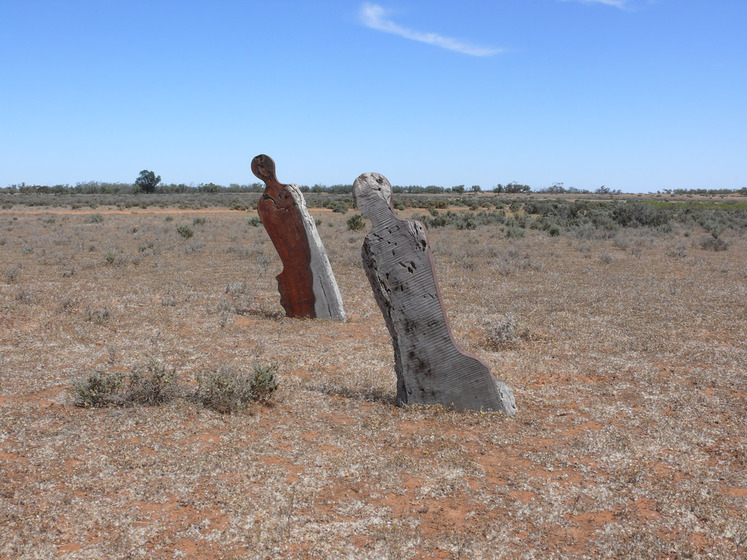Two wooden sculptures, human like in their style, are positioned in an open dry and sandy landscape, the blue sky on the horizon.