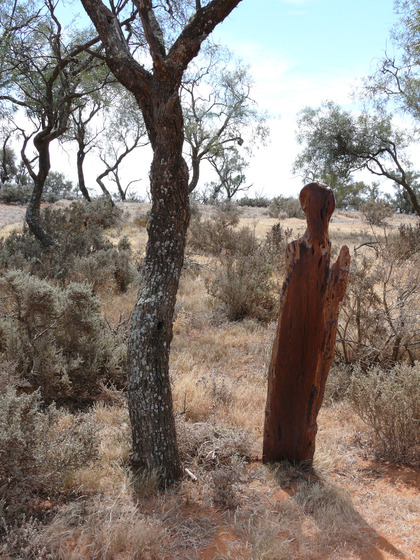 A wooden human like sculpture stands next to a well established tree, lichen growing up the sides. Around the sculpture is lots of bushland scrub and dry grass.
