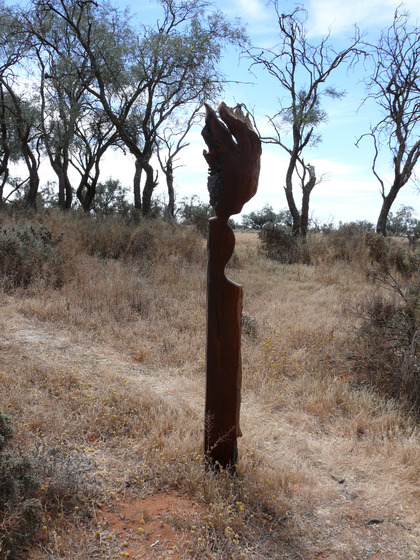 A wooden human like sculpture stands in lots of bushland scrub and dry grass, other taller trees stand in the distance.