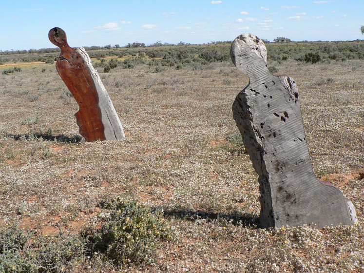 Two wooden sculptures, human like in their style, are positioned in an open dry and sandy landscape, the blue sky on the horizon.