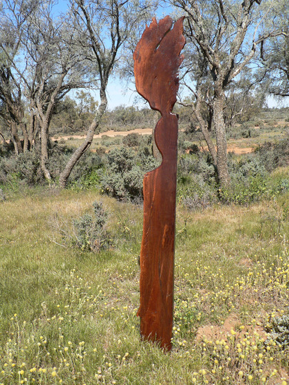 A wooden human like sculpture stands in lots of bushland scrub and dry grass, other taller trees stand in the distance.