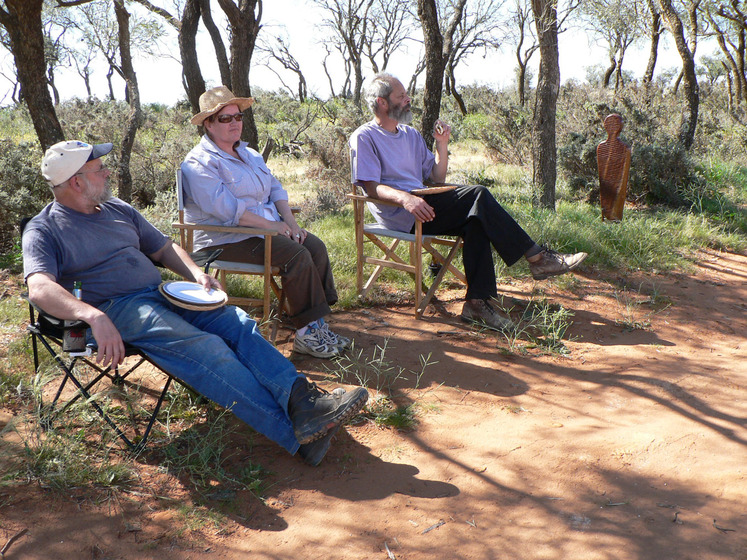 A group of people sit in deck chairs within a shaded patch of bush, the ground red and sandy. Just next to the group is a small wooden sculpture, human like in its form.