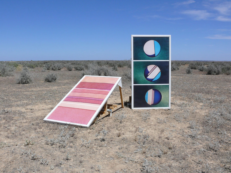 Two large rectangle pieces of card, one leaning on a wooden structure so it looks like a ramp, and another standing straight up right. The leaning piece is coloured in red and yellow stripes and the upright piece is divided into three parts, each with a circle shape filled with greens, pinks and blues. It is set in a dry, dusty and scrubby landscape.