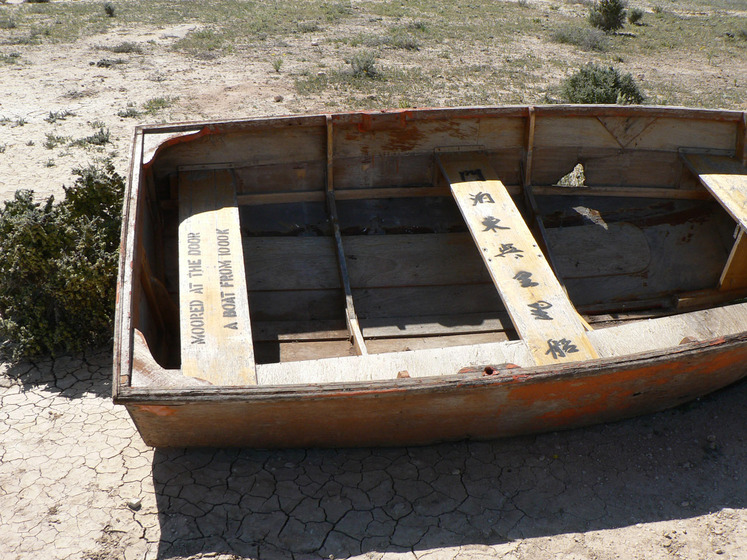 A wooden row boat sits in a dry, dusty and scrubby landscape. On the back and middle wooden seats is printed writing, one english and one chinese characters.