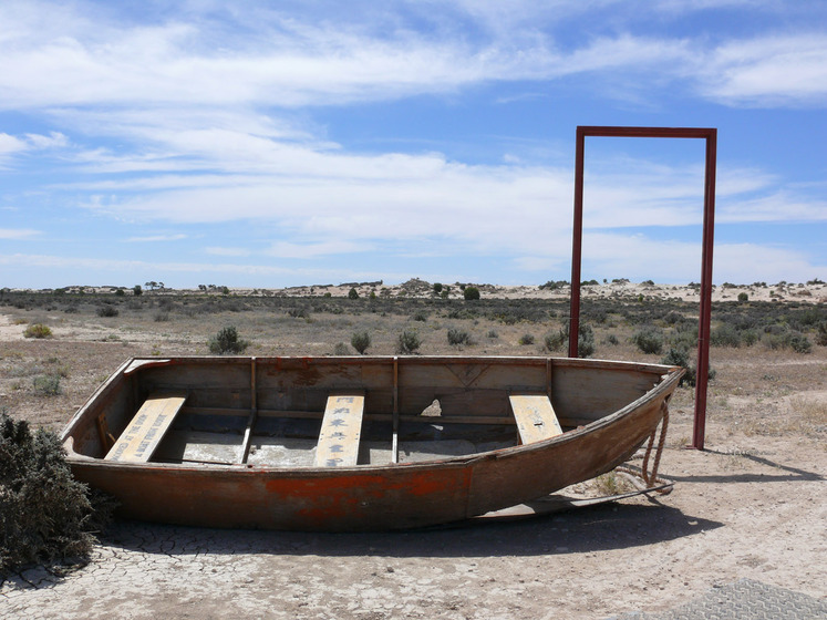A wooden row boat sits in a dry, dusty and scrubby landscape. On the back and middle wooden seats is printed writing, one english and one chinese characters. Behind the boat is a rusty door frame, with no door.
