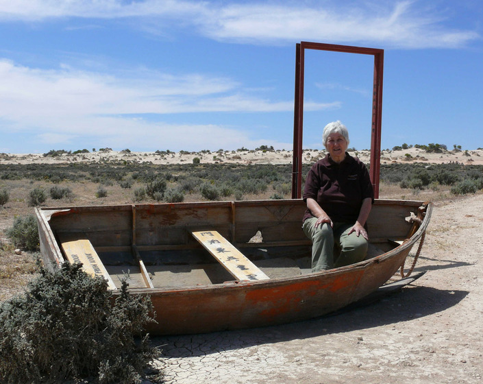 A wooden row boat sits in a dry, dusty and scrubby landscape. On the back and middle wooden seats is printed writing, one english and one chinese characters. Behind the boat is a rusty door frame, with no door. An older woman sits in the boat and smiles at the camera.
