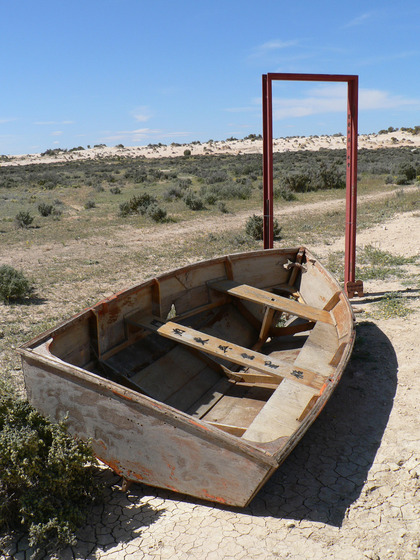 A wooden row boat sits in a dry, dusty and scrubby landscape. On the back and middle wooden seats is printed writing, one english and one chinese characters. Behind the boat is a rusty door frame, with no door.