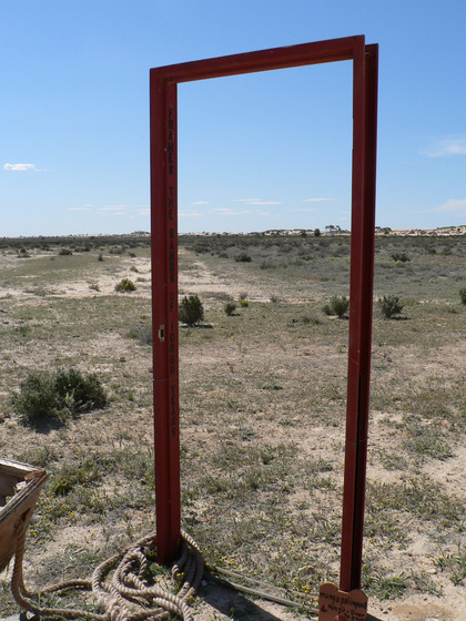 A rusty door frame, with no door, stands in a dry, dusty and scrubby landscape.