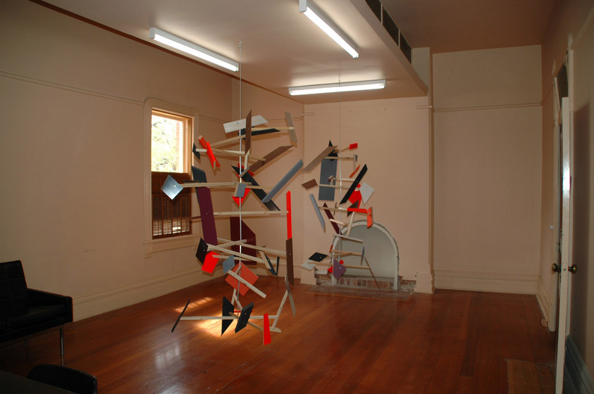 A mobile hangs from the ceiling of a room with wooden floorboards, white walls and a small fireplace at the back. The mobile is made of different coloured shapes, hanging at various angles and attached to intercepting pieces of dowel.