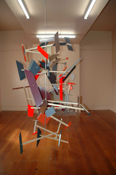 A mobile hangs from the ceiling of a room with wooden floorboards, white walls and a small fireplace at the back. The mobile is made of different coloured shapes, hanging at various angles and attached to intercepting pieces of dowel.