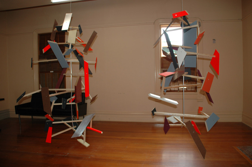 Two mobiles hang from the ceiling of a room with wooden floorboards, white walls and two side by side windows. The mobiles are made of different coloured shapes, hanging at various angles and attached to intercepting pieces of dowel.