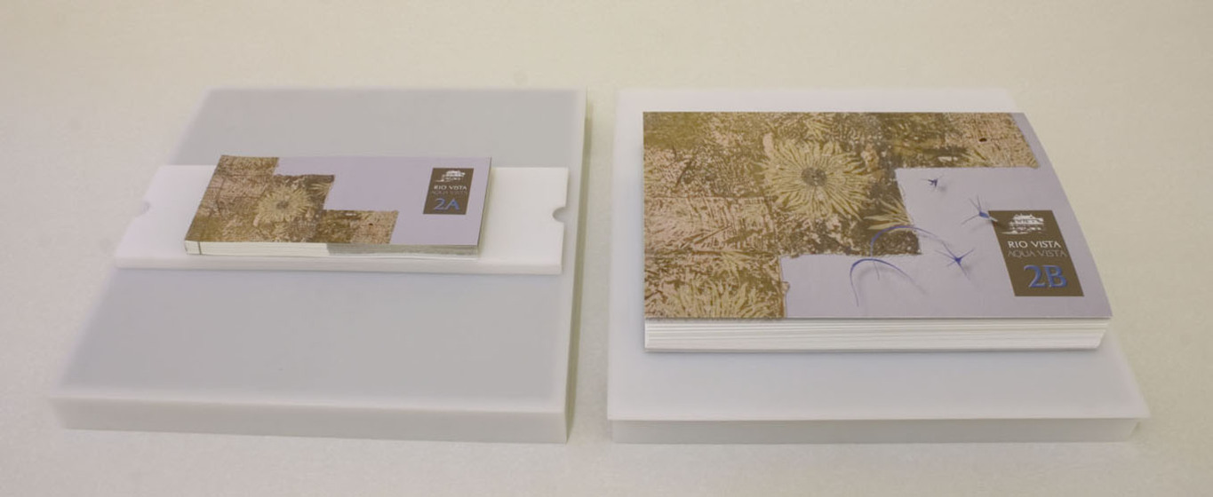 Two artists books, one small and rectangular and the other large and square, sit flat on two white square mounts.