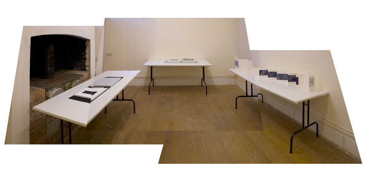 A photograph of an exhibition space, with three trestle tables set up against three walls. One trestle table is positioned front of an old stone kitchen stove, whilst the others are infront of white walls. On each table is an example of a book - some flat and closed, others open and concertinaed out for visibility on all pages.