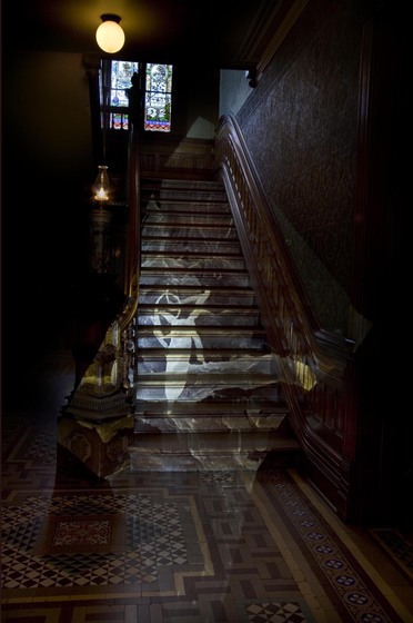 A photograph of a darkened stairwell, ornate tiles on the floor and dark wooden walls surrounding the staircase. At the top of the staircase is a large colourful stain glass window. Projected onto the staircase is a light installation, of curved white shapes.