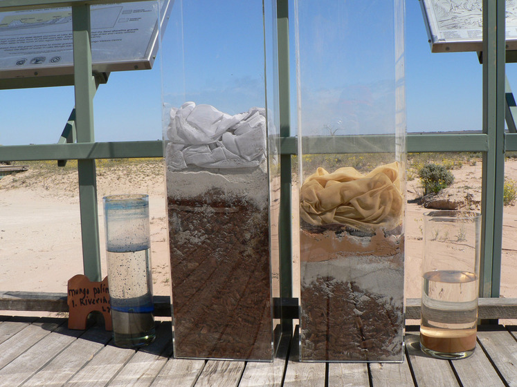 A wooden boardwalk on a sandy beach, holding four glass vessels, each containing water and some other substance, such as sand, dirt and clothing. Information boards can be seen slightly behind the boardwalk.