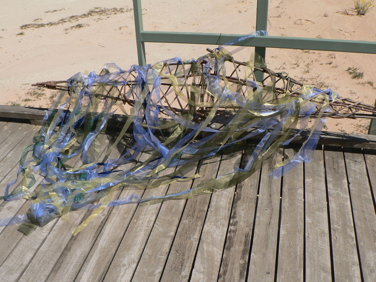 A wooden boardwalk on a sandy beach with a wire mesh shape resting on the top. Attached to the structure are blue and yellow strips of cellophane, blowing slightly.