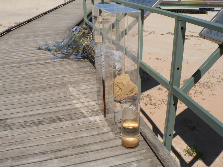 A wooden boardwalk on a sandy beach, holding four glass vessels, each containing water and some other substance, such as sand, dirt and clothing. Next to the vessels is a wire mesh object with blue and yellow cellophane strips attached.