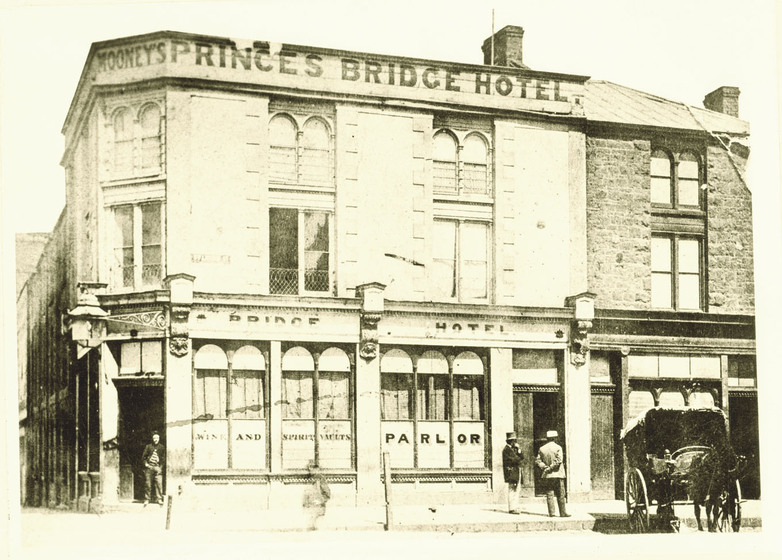 Sepia photograph of a two story brick building on the corner of a street. Outside stand two men next to a horse and cart, while another man leans in the doorway.
