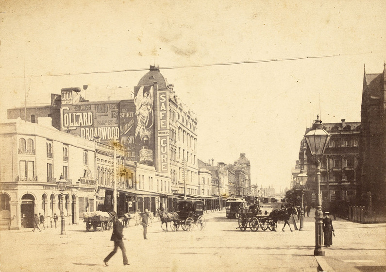 Street scene of a wide intersection with brick buildings of differing heights lining both sides of the street. People walk through the scene and horse and carts turn in various directions on the dirt roads.