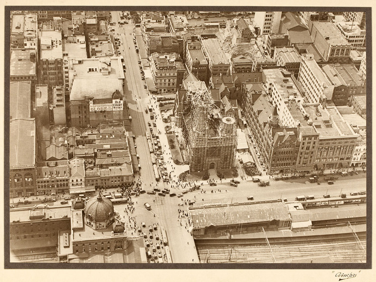Sepia photograph of an aerial view of a busy city intersection with construction on St Paul's Cathedral taking place on one corner. Cars can be seen heading in all four directions of the intersection.