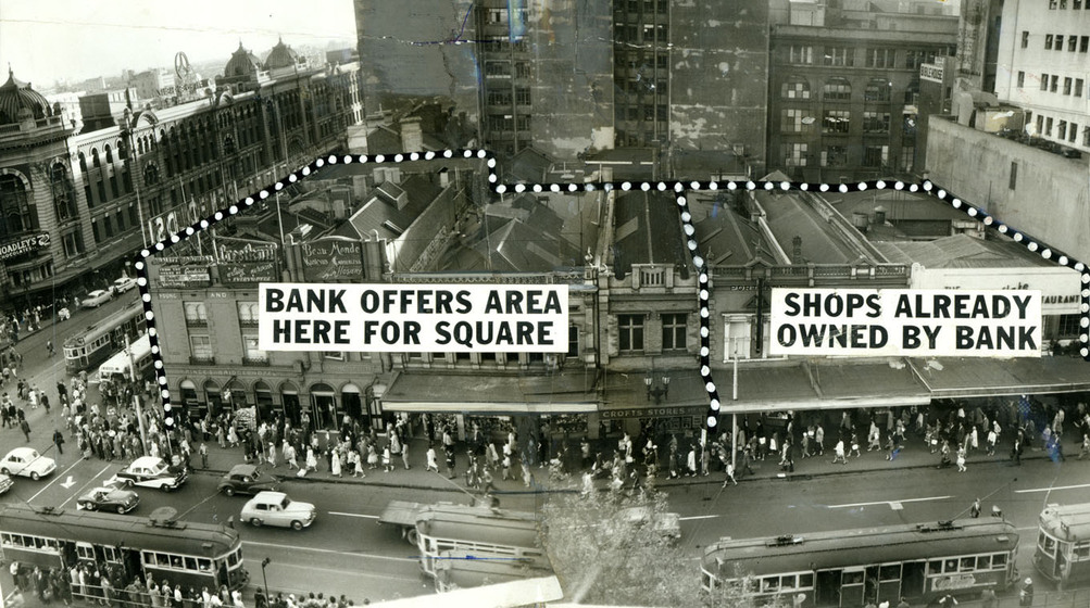 Photograph taken from a tall building looking down on a busy streetscape - lots of pedestrians walk in front of the brick buildings and cars and trams drive down the centre of the road. Captions are placed over the top of the photograph, stating 'Bank offers area here for square' and 'Shops already owned by bank'.