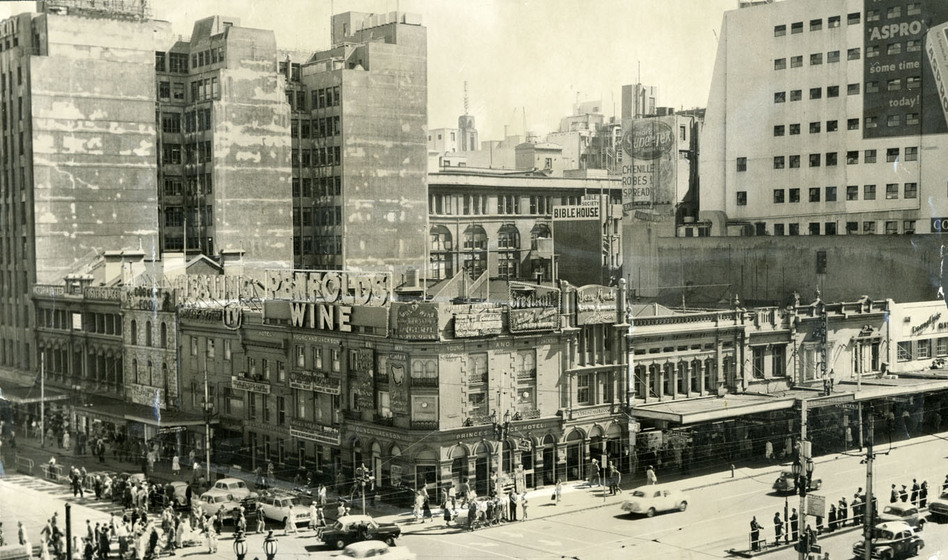 Black and white photograph of a busy streetscape, with small two story brick buildings on the street front and tall glass towers dwarfing them in the background. People walk across the intersection and cars drive down the centre of the road, some stopped at traffic lights.