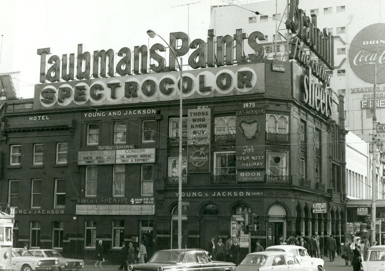 Black and white photograph of a three story brick building on the corner of a busy intersection. Large neon light letters are perched on top, stating ' Taubman's Paints Spectrocolor'.
