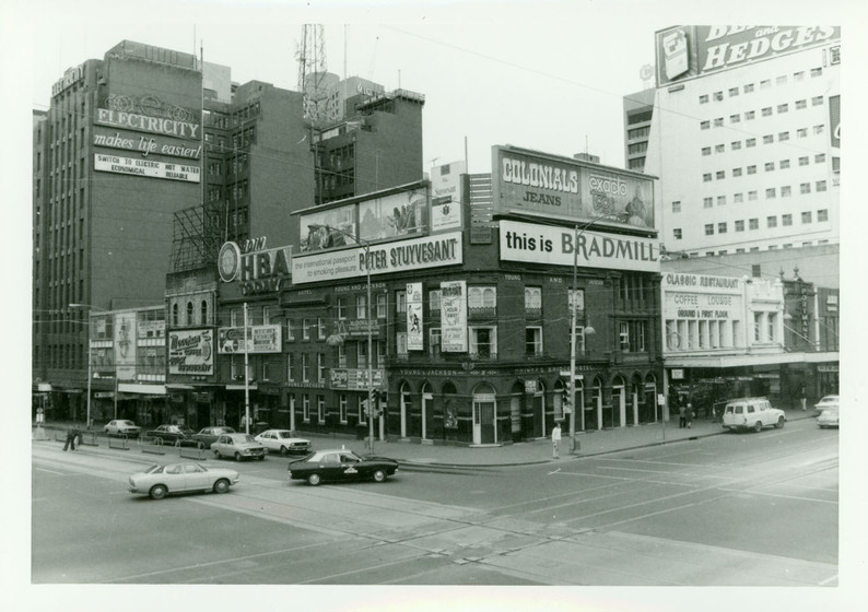 Black and white photograph of a three story brick building on the corner of a busy intersection. Tall skyscrapers stand behind the building, short by comparison, and cars and pedestrians across the intersection in front.