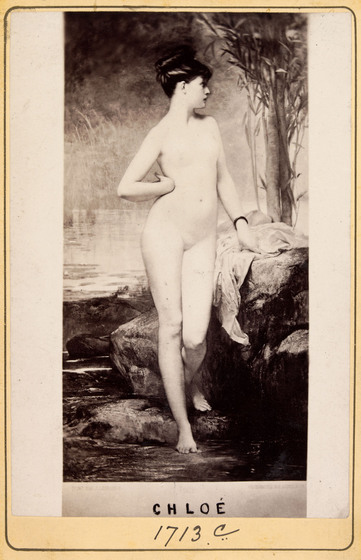 A naked woman stands with one hand on her hip and the resting on a nearby stone. Her face is turned to the side and a painted backdrop of a river and trees stands behind her.