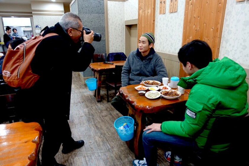Man stands in a restaurant with a long lense camera in his hand, up to his eye. He is photographing two men sitting at a table with plates of food in front of them.