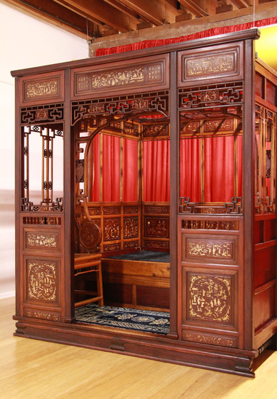 An ornate wooden structure resembling a tall box with an open doorway and windows. Red curtains are positioned in front of some of the window cut outs and gold images decorate the wood panels on the front. Within the structure is a bed, with green lining where a mattress would go, a wooden chair to the side and a carpert on the floor. More decorative motifs line the wooden inside of the structure.
