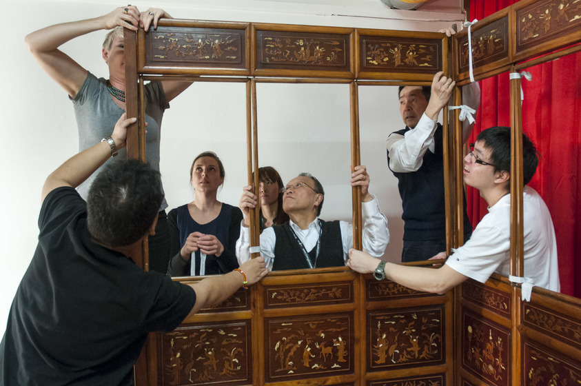 A group of seven stand around a wooden structure - some have their hands on the wood, holding it upright - their hands in various positions. Some standby staring at the people holding the structure, observing intensely what is being done.