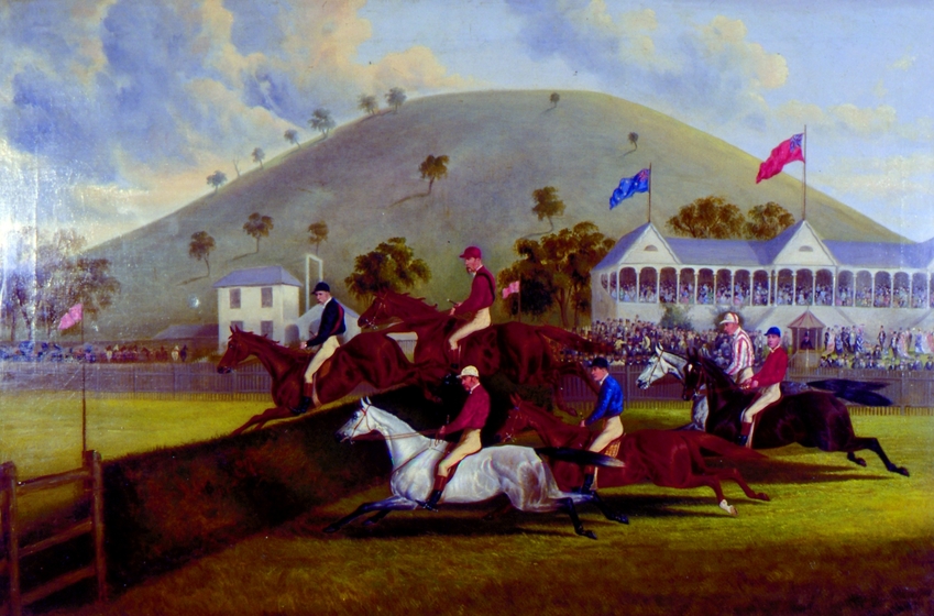 Six horses of different heights and colours, all with jockeys on board, ride towards a grass jump on a green race track. In the background are large white spectator stands with red and blue flags waving on top. Beyond that is a grassy mound with palm like trees across the surface.