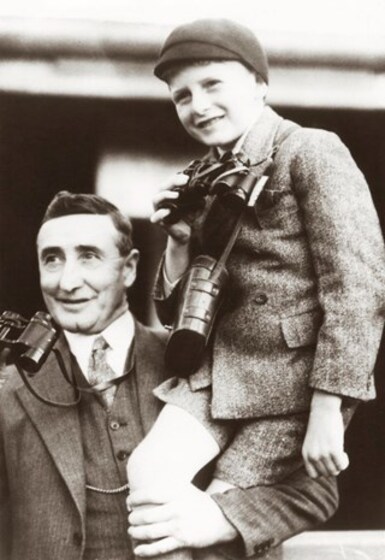 An older man in a suit and tie holds a young boy, also in a suit, up on his shoulder. Both are holding binouclars in their right hand, held halfway up towards their face - they are both smiling.