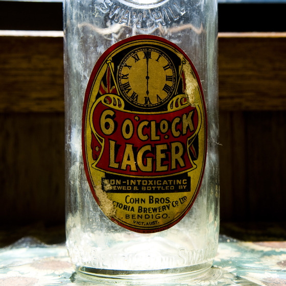 Clear glass bottle with a gold, red and black label, including a picture of a clock with the hands set at 6 o'clock.