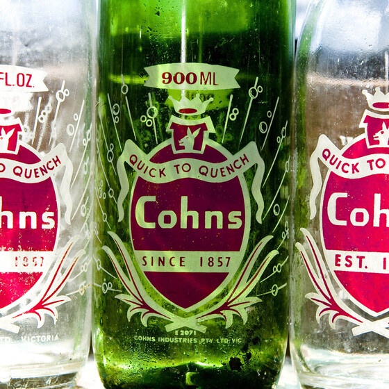 Three glass bottles in a row, two clear and the one in the middle green glass. They all have labels in red and white colouring and the saying 'quick to quench'.